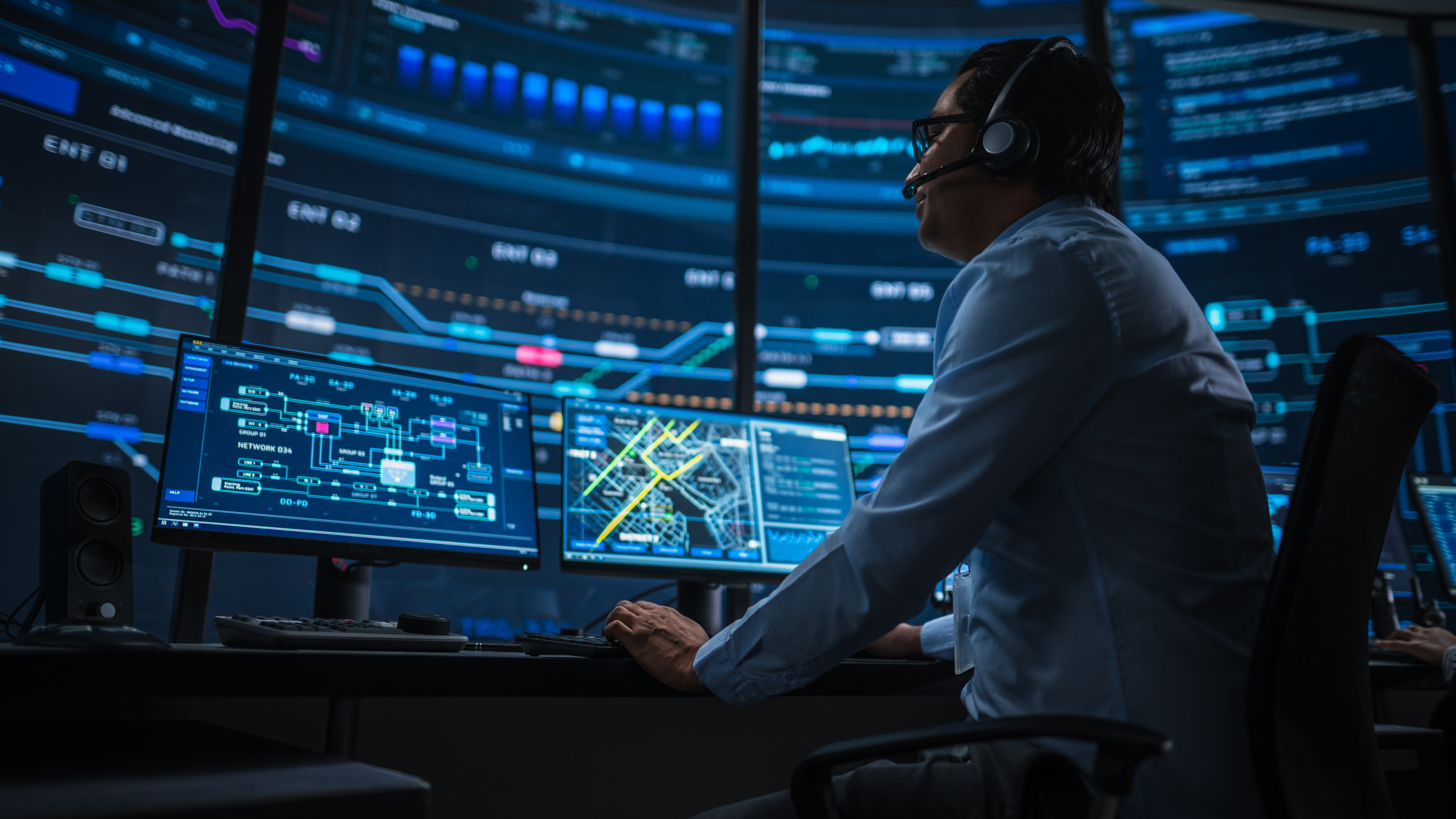 Professional IT Technical Support Specialist and Software Developer Working on Computer in Monitoring Control Room with Digital Screens. Employee Uses Headphones with Mic and Talking on a Call.